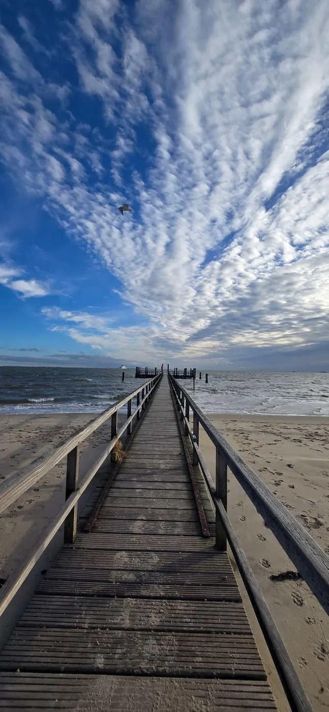 Hölzerner Badesteg führt am Strand von Föhr ins Meer, darüber ein weiter Wolkenhimmel